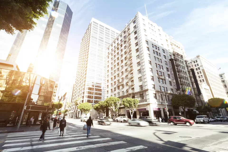 Intersection in downtown Los Angeles with pedestrians crossing, vintage building across street, and modern glass office tower in background.